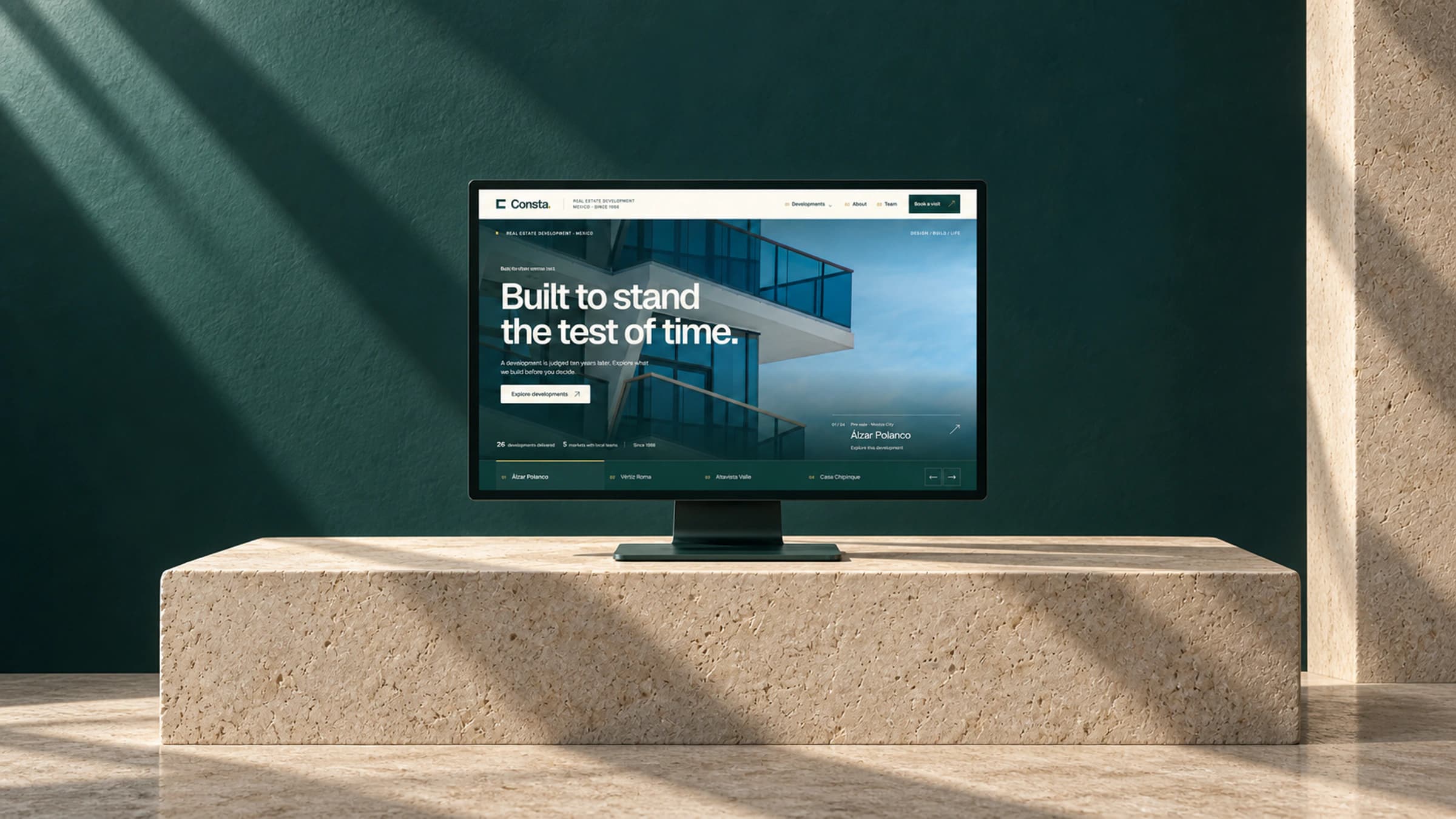 Consta’s panoramic homepage on a dark monitor, with a limestone plinth and a continuous petrol-green architectural backdrop.